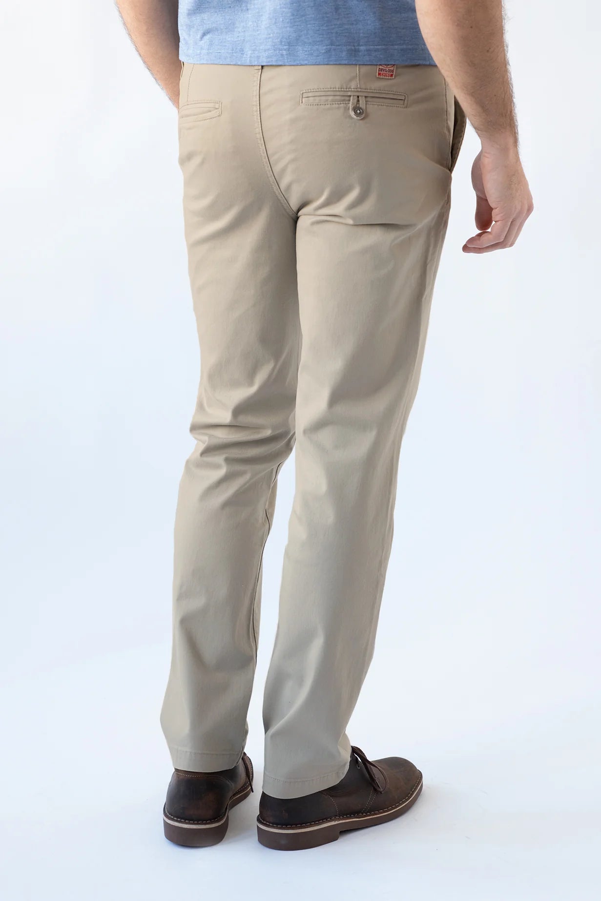 A man viewed from the back wearing beige slim-fit chino pants, a light blue t-shirt, and dark brown leather loafers, set against a plain white studio background.