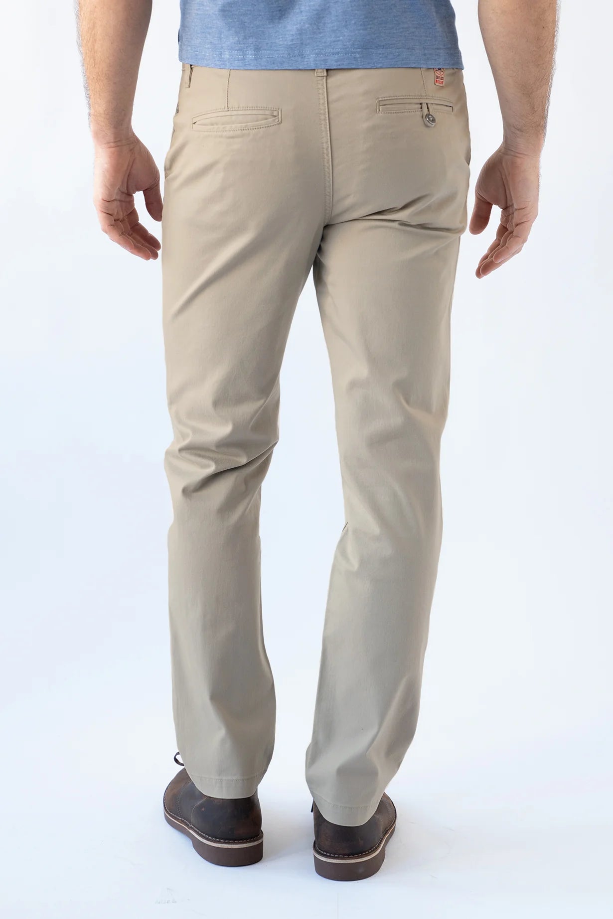 A person shown from the back wearing light beige chino pants, a blue shirt, and dark brown shoes against a plain white studio background.