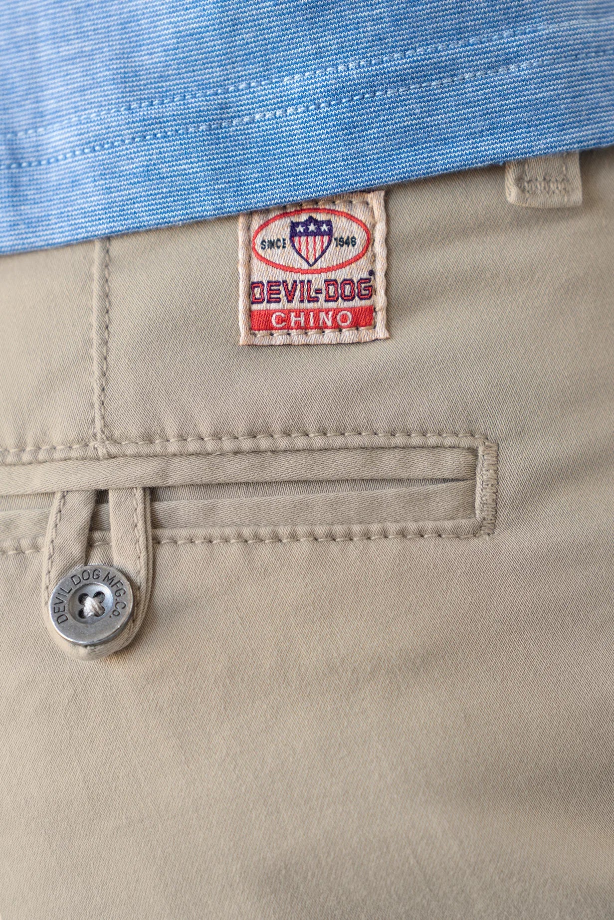 Close-up of khaki chino pants showing a Devil Dog Chino brand patch, a buttoned welt pocket, and a blue striped shirt tucked into the waistband.
