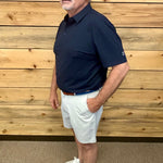 A man wearing a navy blue short-sleeve polo shirt, white shorts, a brown belt, and white sneakers, standing in front of a light wood plank wall.