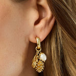 A close-up of a model's ear wearing a gold huggie hoop earring with dangling gold leaf, pinecone, and white pearl charms.
