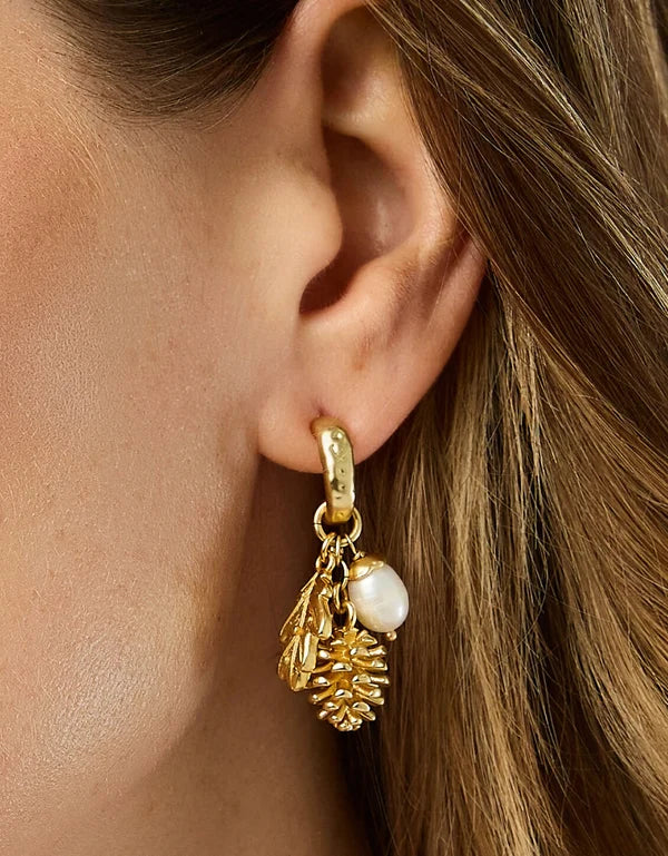 A close-up of a model's ear wearing a gold huggie hoop earring with dangling gold leaf, pinecone, and white pearl charms.