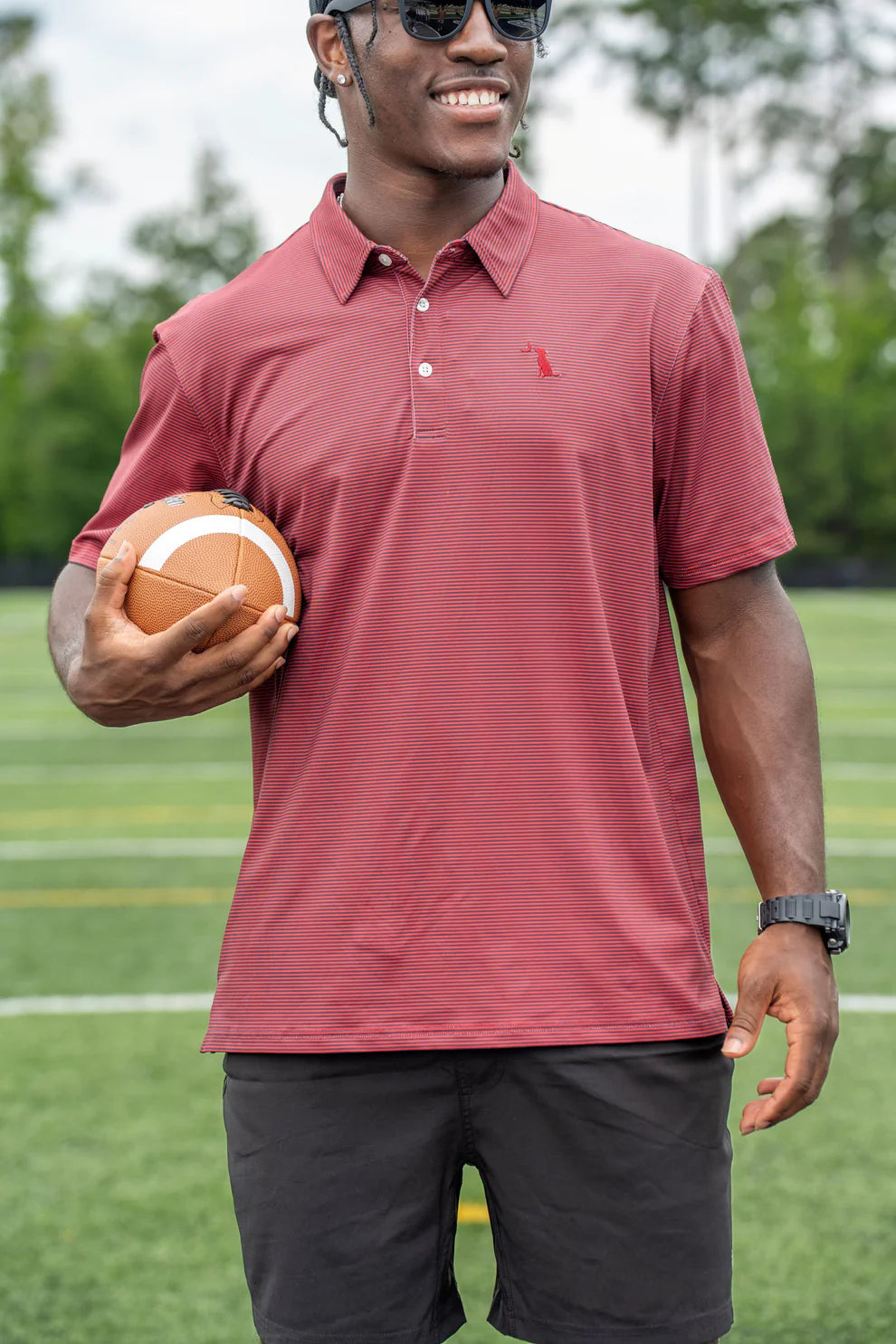 A man wearing a muted red polo shirt and black shorts stands on a green sports field holding a football.