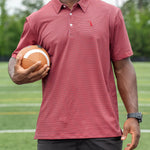 A man wearing a muted red polo shirt and black shorts stands on a green sports field holding a football.