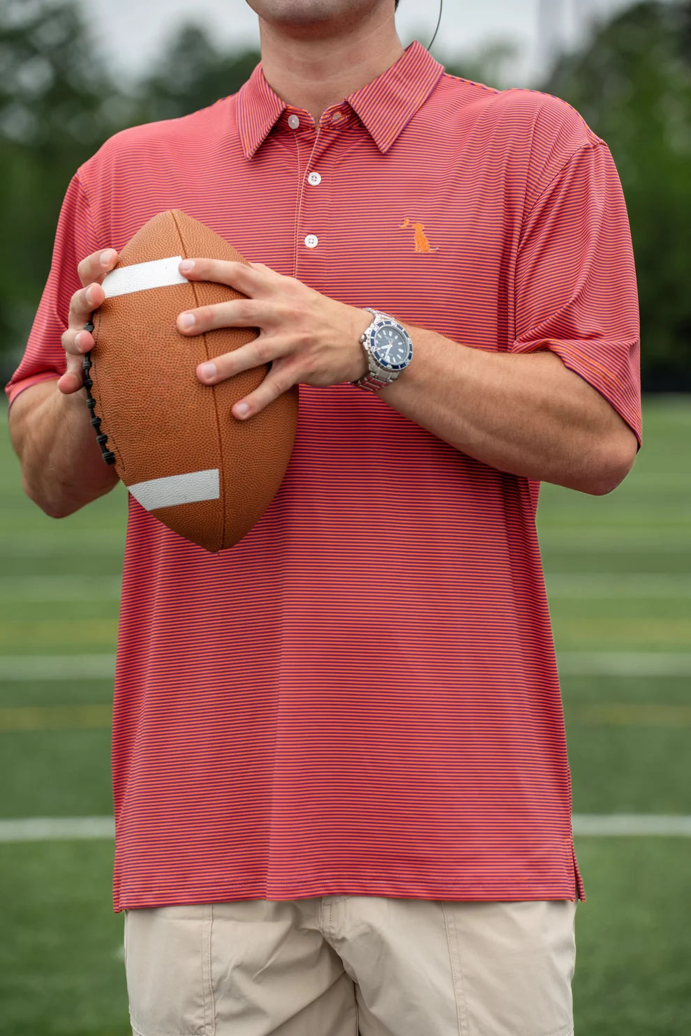 A man wearing a coral red polo shirt with a small orange embroidered logo on the chest, holding a brown football on a green field.