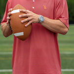 A man wearing a coral red polo shirt with a small orange embroidered logo on the chest, holding a brown football on a green field.