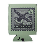 A sage green can cooler featuring a vintage-style duck stamp design with a goose, a duck, and the text 'LOCAL BOY OUTFITTERS'. The bottom tab of the can cooler has a black dog silhouette and a crescent moon, set against a plain white background.