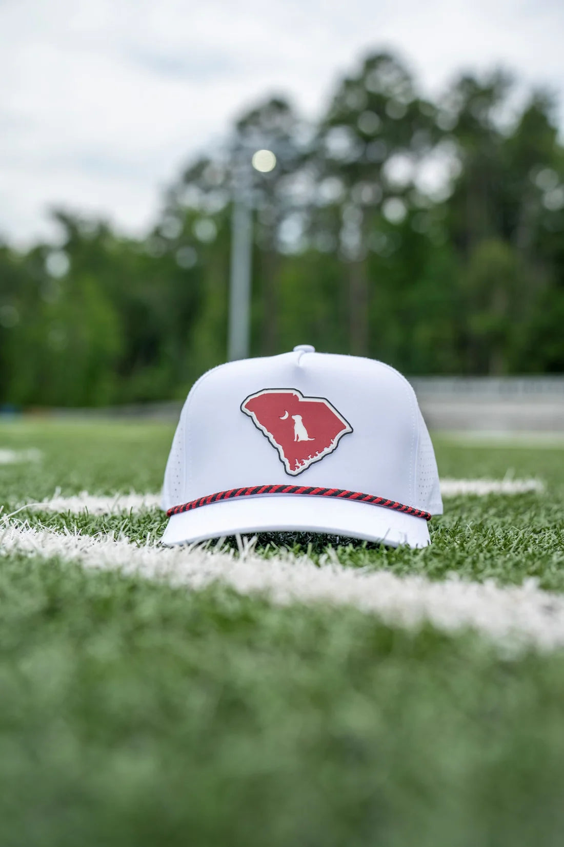 A white baseball cap with a red logo on the front, resting on a green athletic field with blurred trees in the background.