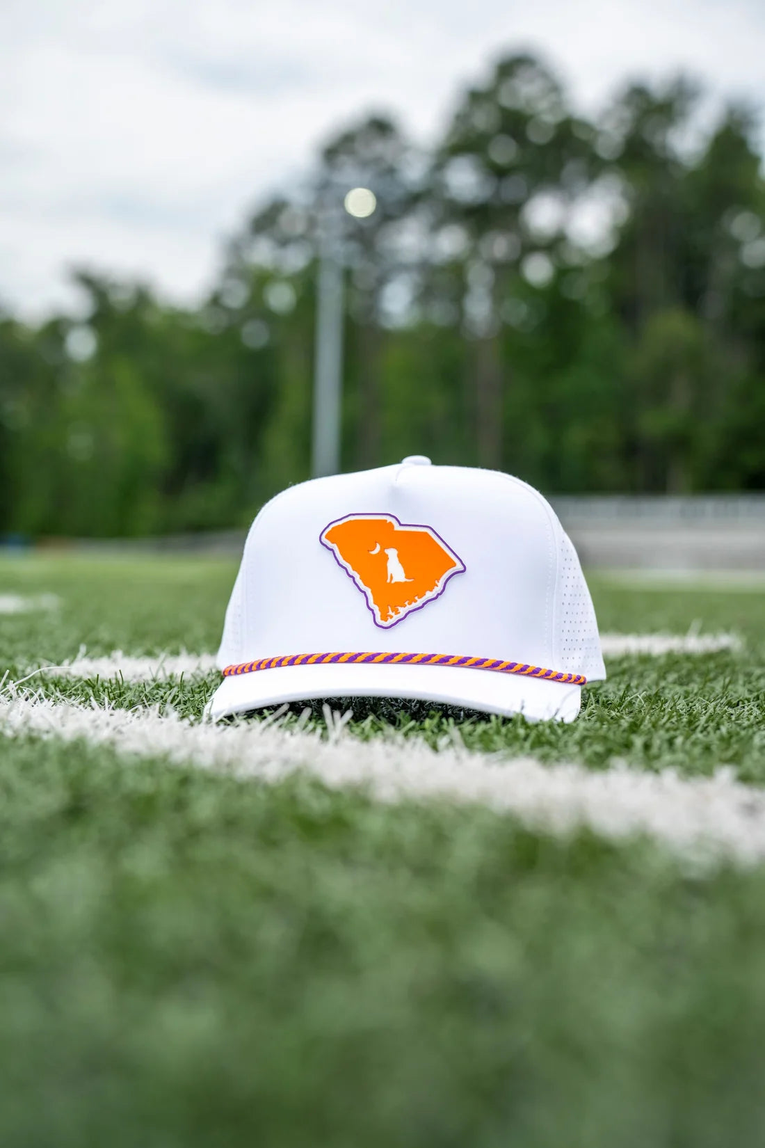 A white trucker-style baseball cap with an orange South Carolina logo patch, resting on a green grass field with white yard lines and a blurred background of trees and stadium lights.