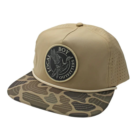 A tan baseball cap with a flat brim featuring a brown and beige topographic map pattern, and a circular black patch on the front that reads 'LOCAL BOY OUTFITTERS' with a bird design.