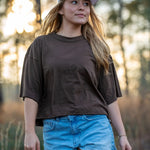 A woman wearing a brown oversized t-shirt, light blue denim shorts, and a blue baseball cap with 'LOCAL' printed on the front, standing outdoors in a wooded setting.