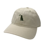 A beige baseball cap featuring a small dark green embroidered graphic of a sitting dog and a crescent moon on the front panel, shown against a plain white background.