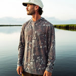 A man wearing a long-sleeve camouflage-patterned fishing shirt and a white cap stands by a calm lake at sunset.