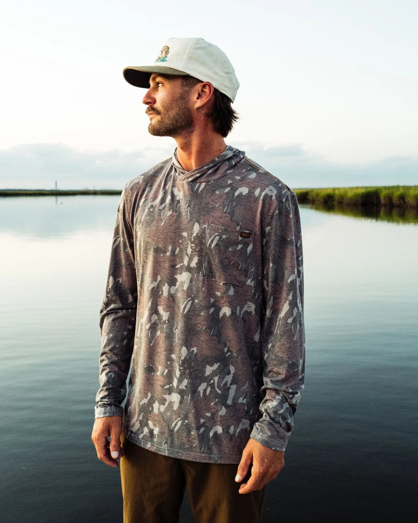 A man wearing a long-sleeve camouflage-patterned fishing shirt and a white cap stands by a calm lake at sunset.
