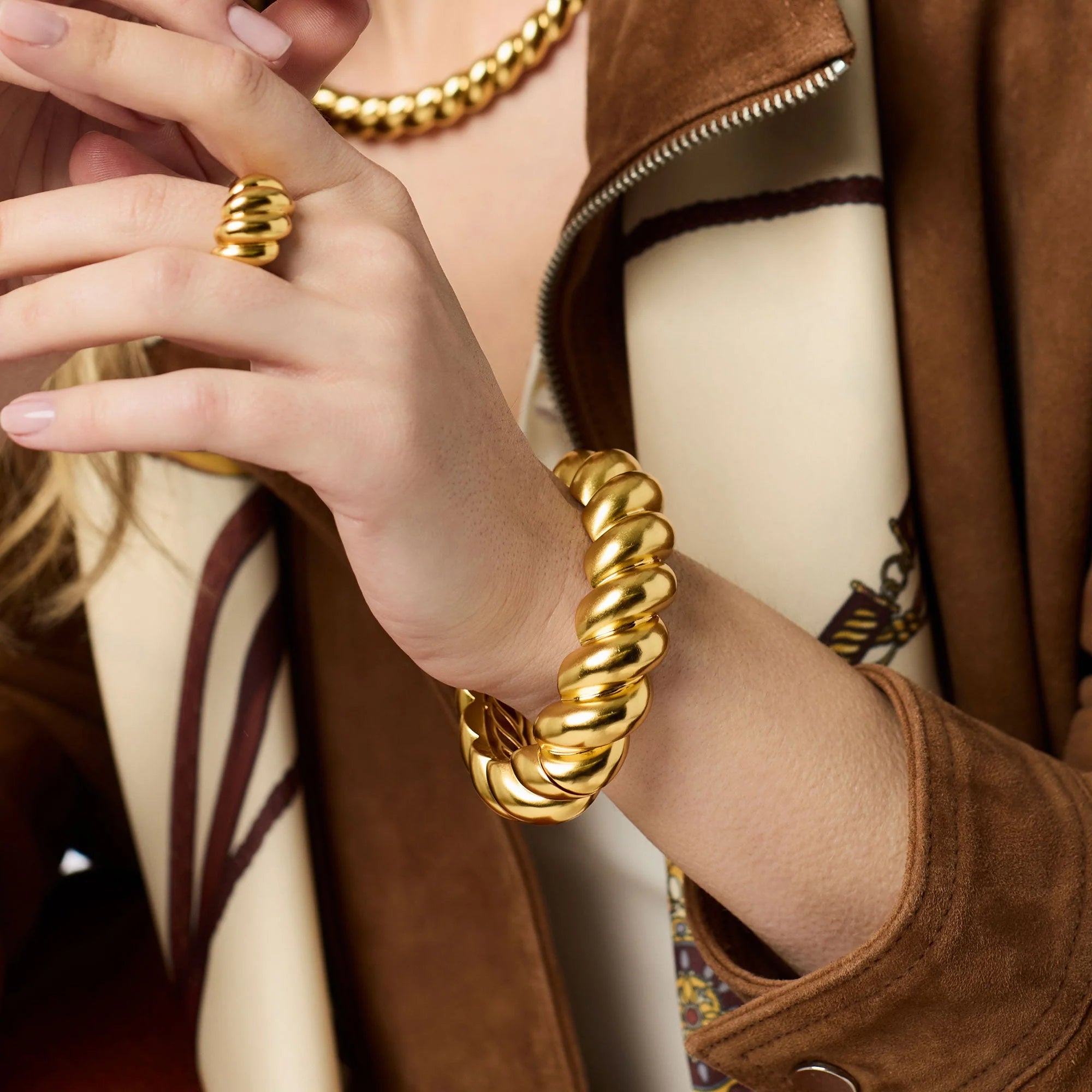 Close-up of a person wearing a gold bracelet and ring, with a brown coat and scarf in the background.