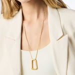Gold necklace with a rectangular pendant worn by a person in a cream blazer.