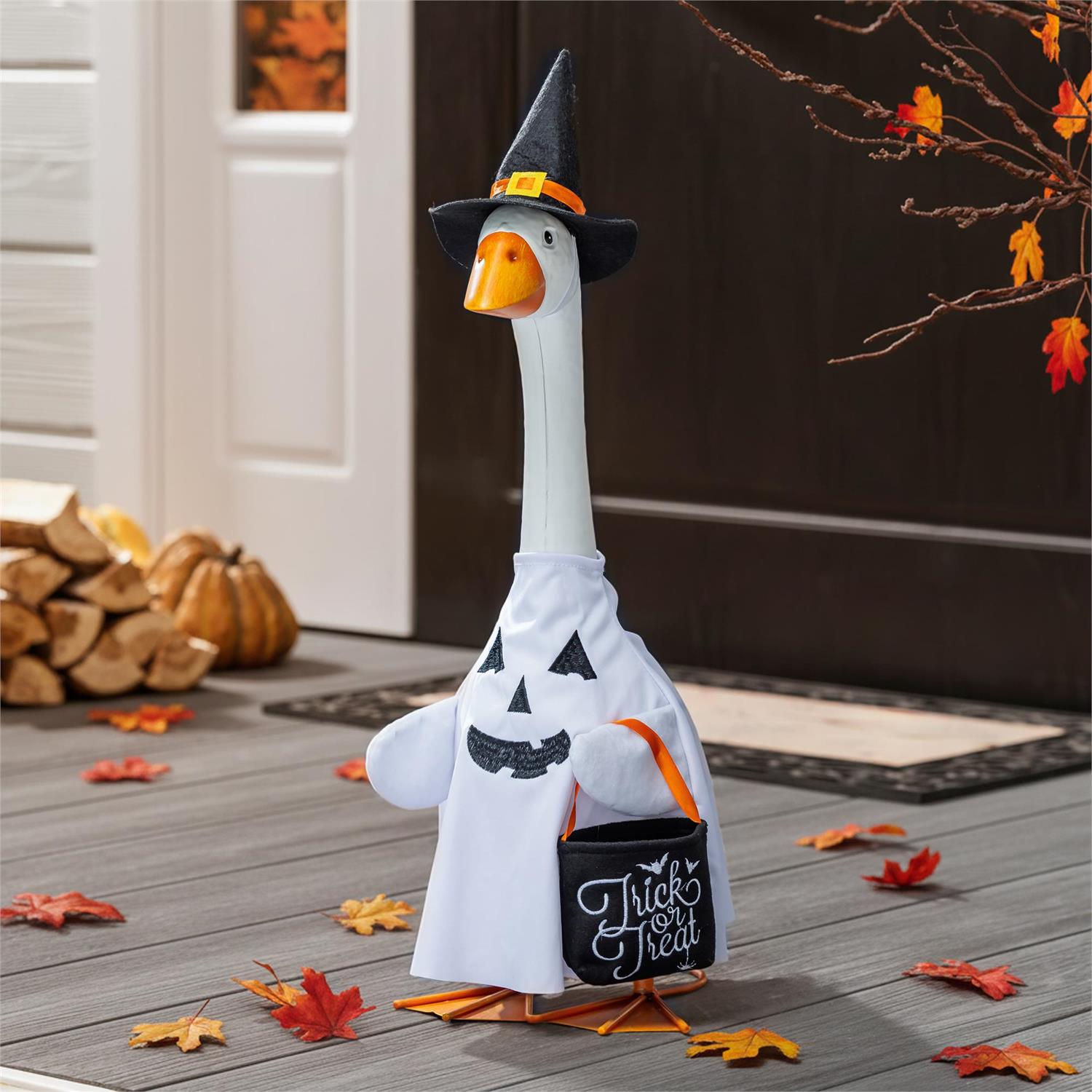 A decorative goose-shaped Halloween decoration wearing a black witch hat and white ghost costume, holding a small black bucket that says "Treat" in script. The scene is staged on a gray porch floor with autumn leaves, pumpkins, stacked firewood, and a dark door in the background.