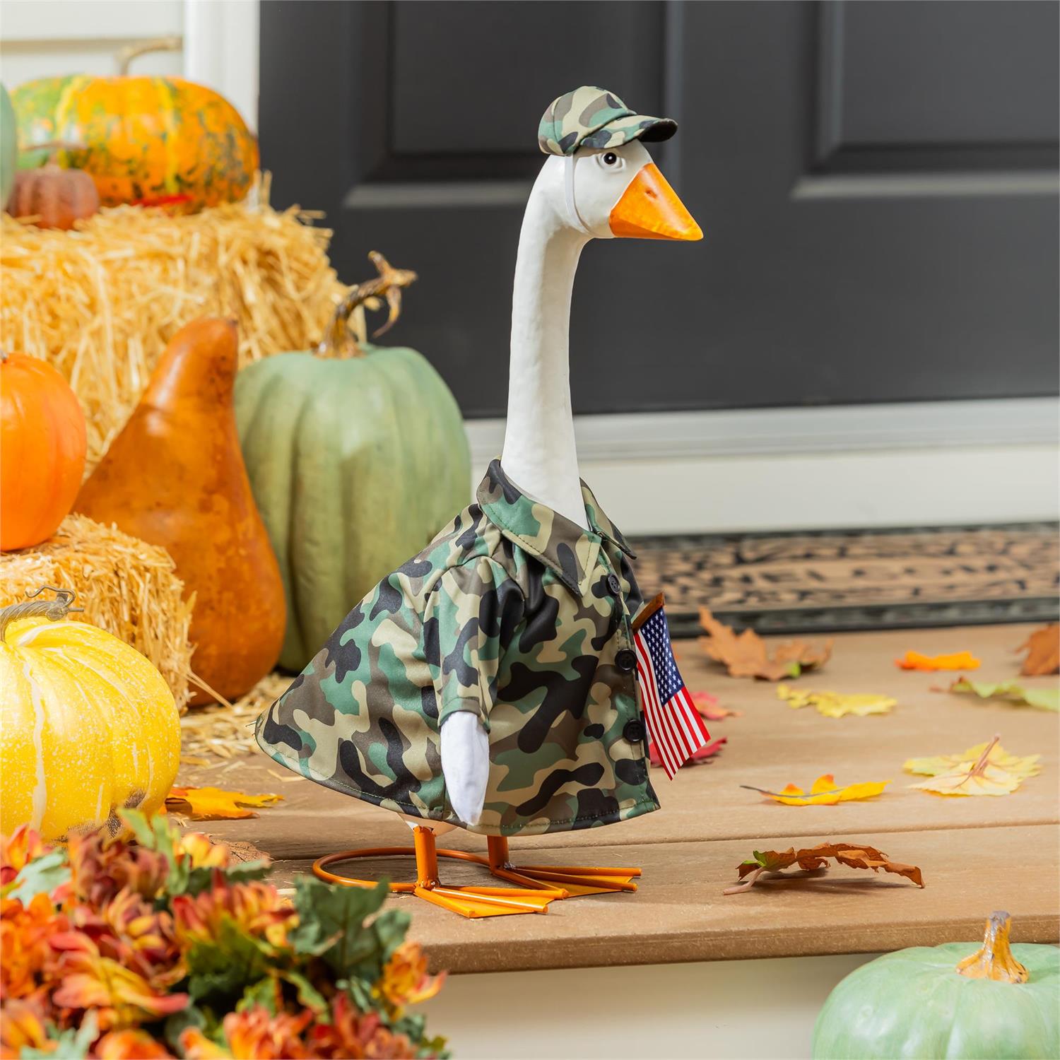 Giddy the Goose wearing a camouflage outfit with matching camo hat, displayed on a fall porch with pumpkins and autumn décor.