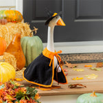 Giddy the Goose wearing a witch costume with black cape, pointed hat, and broom, displayed on a fall porch with pumpkins and Halloween décor.