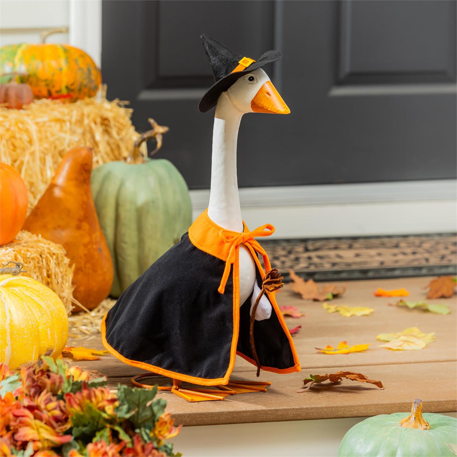 Giddy the Goose wearing a witch costume with black cape, pointed hat, and broom, displayed on a fall porch with pumpkins and Halloween décor.