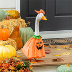 A decorative goose figurine wearing an orange pumpkin dress with a black jack-o'-lantern face and a small orange hat, displayed in a fall-themed setting with pumpkins, hay, leaves, and a dark front door background.