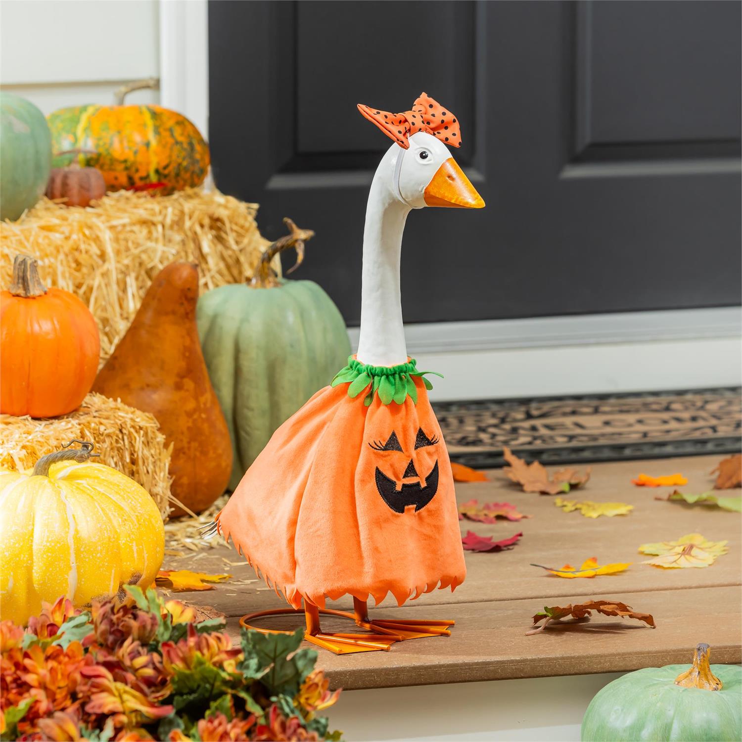 A decorative goose figurine wearing an orange pumpkin dress with a black jack-o'-lantern face and a small orange hat, displayed in a fall-themed setting with pumpkins, hay, leaves, and a dark front door background.
