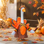 A decorative turkey-shaped garden ornament stands on a wooden deck surrounded by autumn props, including pumpkins, corn, dried grass, and fallen leaves. The ornament has a white neck, orange beak, brown hat, orange and brown body, and orange, yellow, and black feather details.
