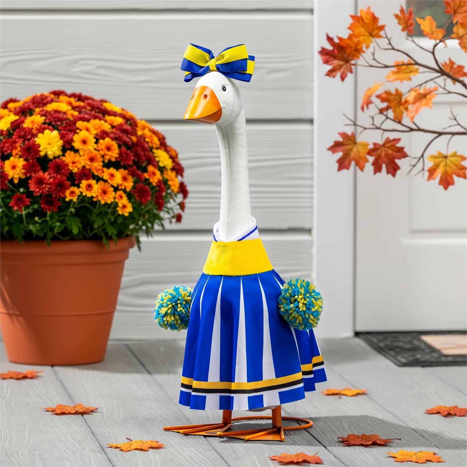 Giddy the Goose dressed as a cheerleader in a blue and gold uniform with matching bow and pom-poms, displayed on a front porch with colorful fall mums.