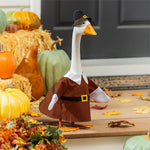Giddy the Goose wearing a Harvest Male Pilgrim Outfit with a brown tunic and black Pilgrim hat, displayed on a fall porch with pumpkins and hay bales.