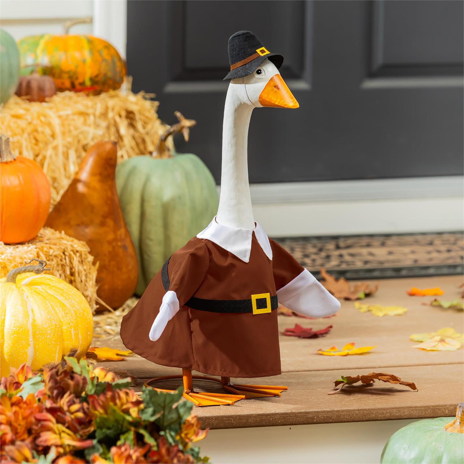 Giddy the Goose wearing a Harvest Male Pilgrim Outfit with a brown tunic and black Pilgrim hat, displayed on a fall porch with pumpkins and hay bales.