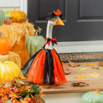 Giddy the Goose wearing a black and orange Witch Outfit with a tulle skirt and witch hat, displayed on a fall porch surrounded by pumpkins and autumn décor.