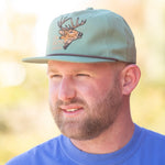 Man wearing a light blue cap with a deer emblem against a blurred natural background