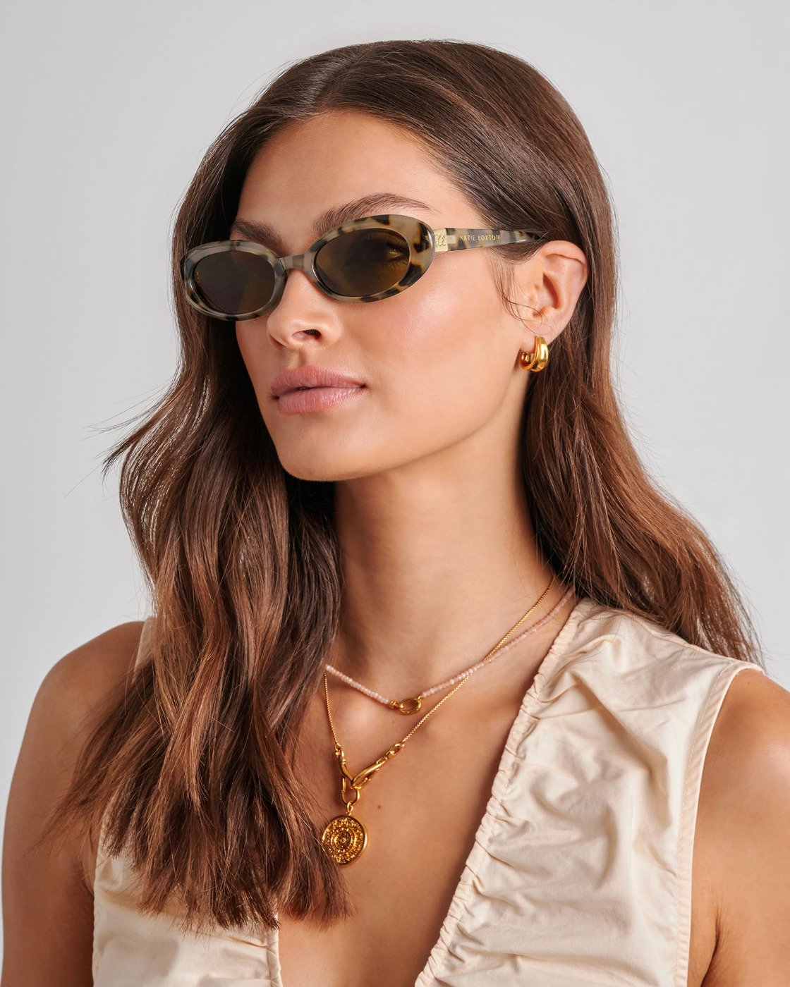 Woman wearing sunglasses and gold jewelry against a neutral background