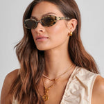 Woman wearing sunglasses and gold jewelry against a neutral background