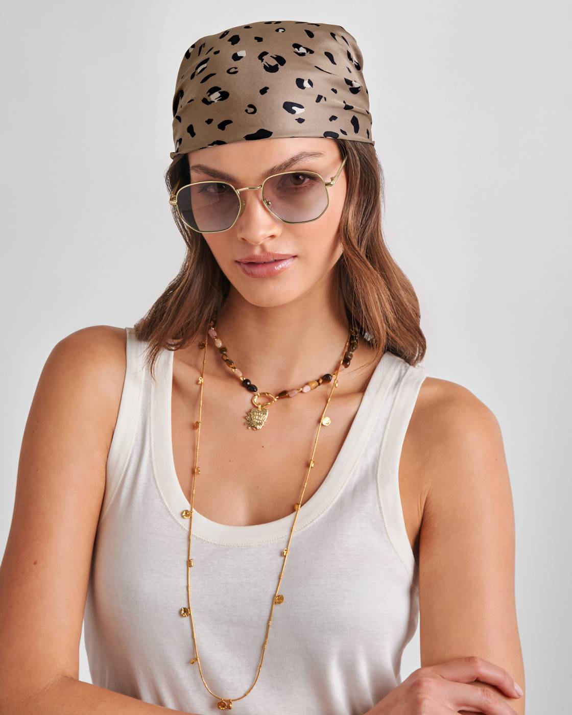 Woman wearing a leopard print headband, sunglasses, and a white tank top with a gold necklace on a light gray background.