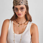 Woman wearing a leopard print headband, sunglasses, and a white tank top with a gold necklace on a light gray background.