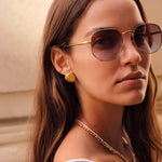 A close-up portrait of a woman wearing gold-framed aviator-style sunglasses with dark tinted lenses, along with gold jewelry, against a softly blurred beige architectural background.