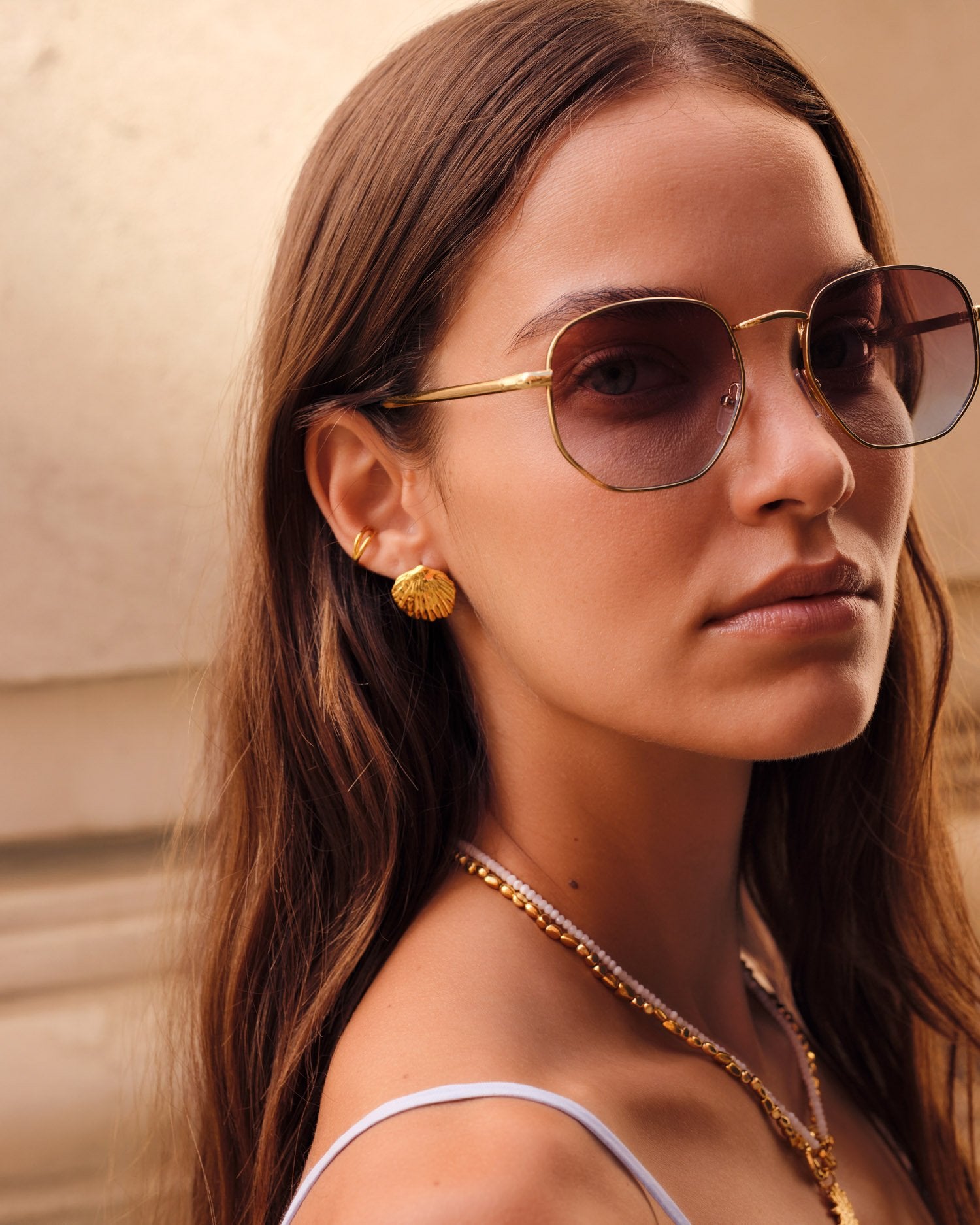 A close-up portrait of a woman wearing gold-framed aviator-style sunglasses with dark tinted lenses, along with gold jewelry, against a softly blurred beige architectural background.