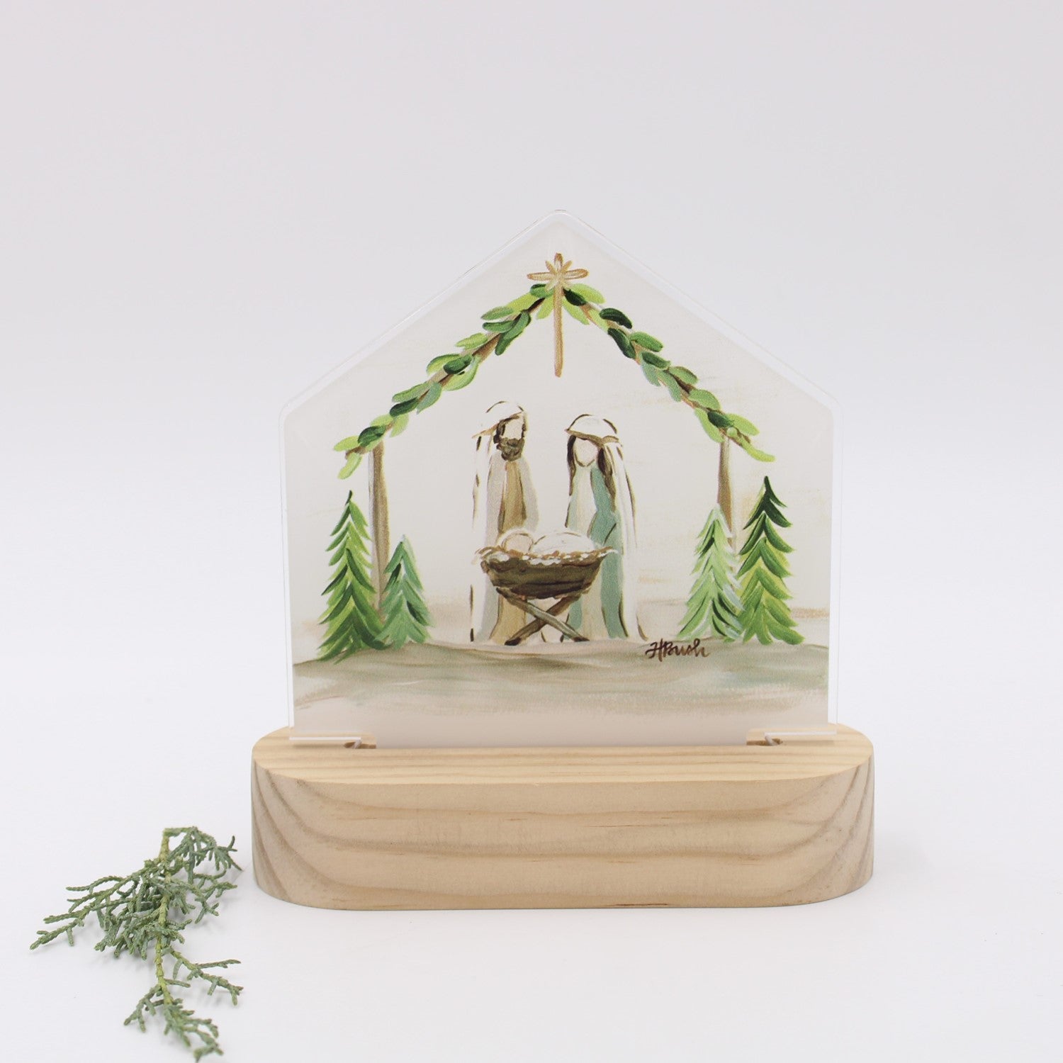 A hand-painted resin nativity scene housed in a clear acrylic house with a light natural wood base, displayed on a clean white background with a small sprig of greenery in the foreground.