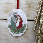 An oval Christmas ornament featuring a painted illustration of Mary and baby Jesus surrounded by a green wreath with red berries, hanging from a red ribbon against a light wood background.